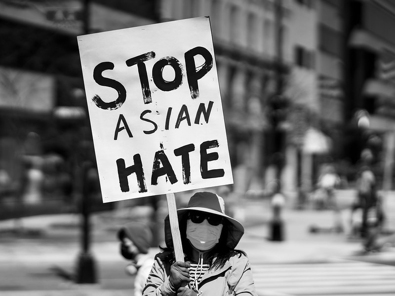 person wearing a surgical mask, sunglasses, and hat holding a sign that says "Stop Asian Hate"